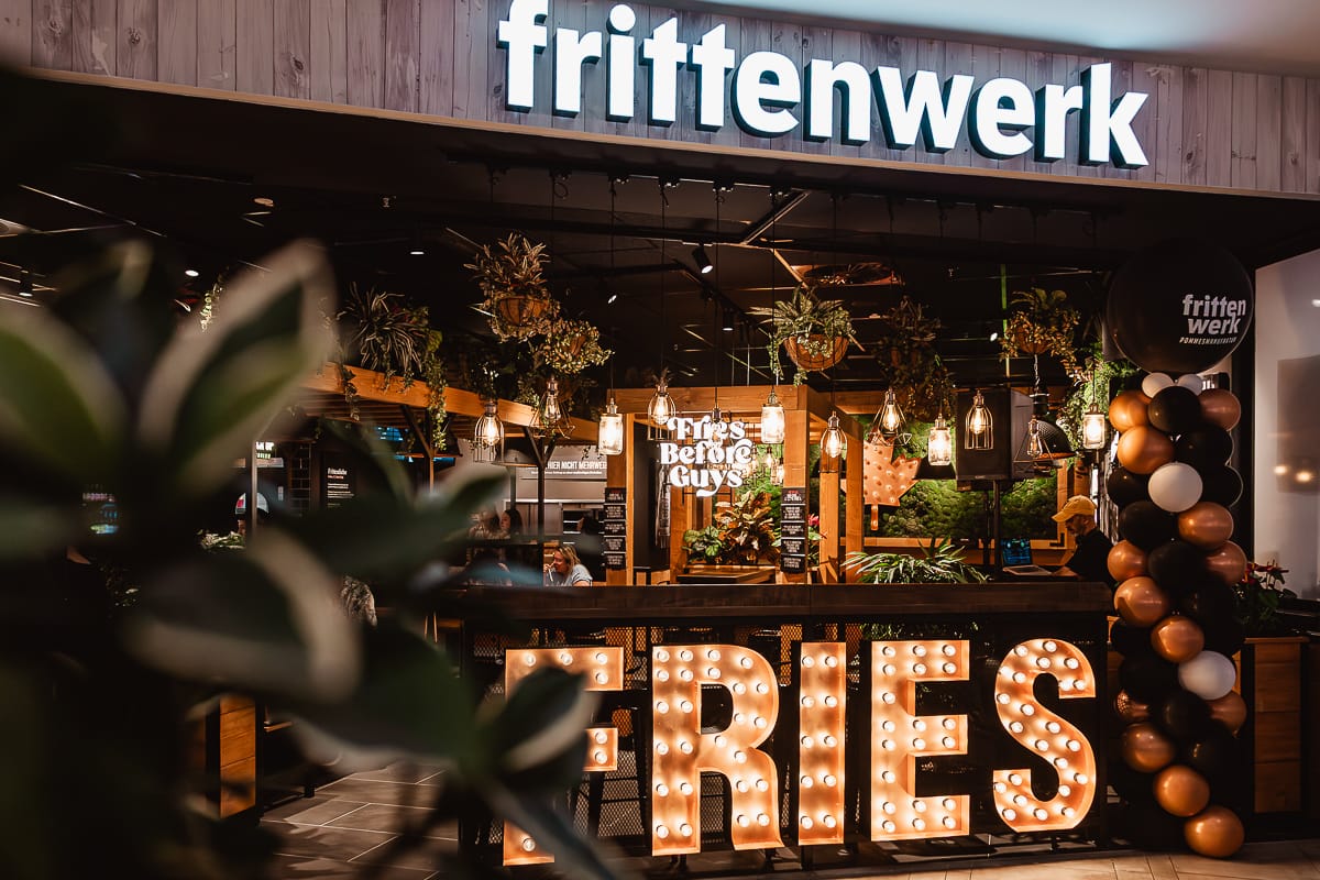 Das Restaurant Frittenwerk Bielefeld hat ein leuchtendes "FRIES"-Schild am Eingang, braune und weiße Luftballons und Blattpflanzen. Im Inneren sorgen eine warme Beleuchtung und hängende Grünpflanzen für eine gemütliche Atmosphäre, und ein "Fries Before Guys"-Schild ist zu sehen.