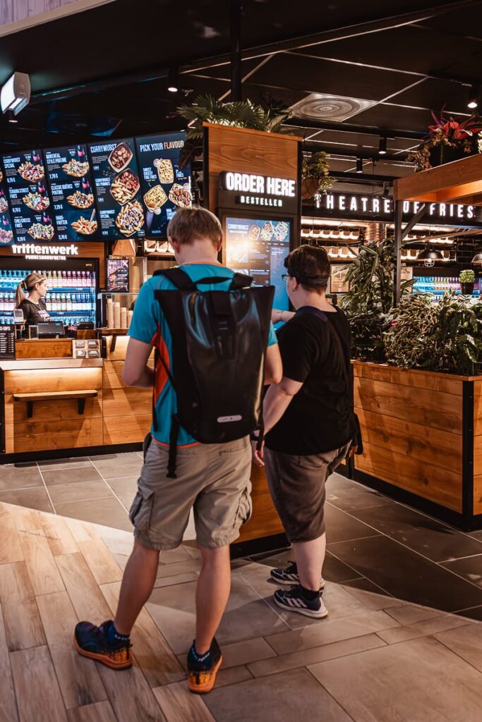 Zwei Personen stehen vor der Fast-Food-Theke von Frittenwerk Bielefeld an einem Bestellterminal, dahinter die Theke. Über der Theke sind digitale Menütafeln angebracht. Die Person links trägt ein blaues Hemd, khakifarbene Shorts und einen großen schwarzen Rucksack, während die andere Person schwarz gekleidet ist. Hinter der Theke wartet eine Mitarbeiterin.