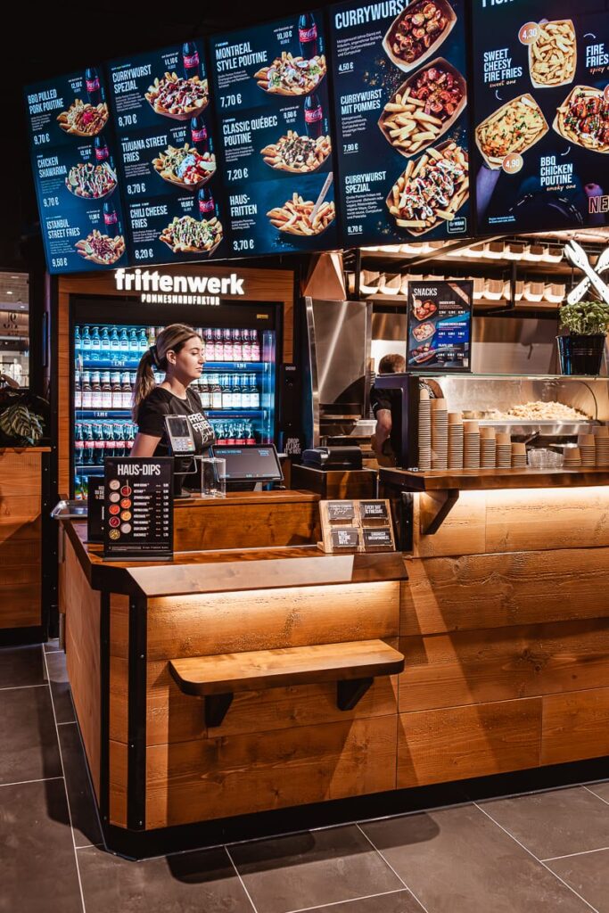 Eine Frau steht hinter dem hölzernen Tresen von Frittenwerk Bielefeld, einem modernen Fastfood-Restaurant. Über ihr zeigen beleuchtete Menütafeln bunte Bilder von Pommes frites, Poutine und Soßen. Hinter ihr sind gekühlte Getränke im Regal zu sehen.