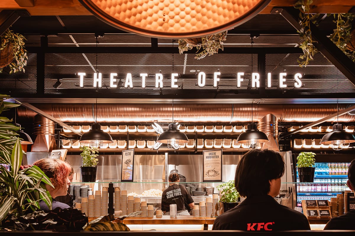 Das Frittenwerk Bielefeld ist ein modernes Lokal mit einer hellen Leuchtreklame mit der Aufschrift "THEATER OF FRIES" über der Theke. Das Personal bereitet das Essen vor, die Kunden warten, es gibt hängende Pflanzen, warmes Licht und Reihen mit verpackten Waren und Getränken.