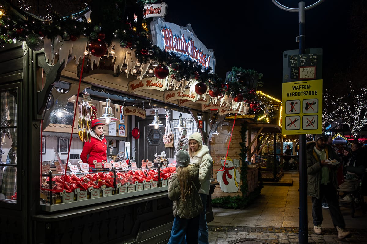 Ein festlicher Weihnachtsmarktstand in der Nacht zeigt rot verpackte Waren und hängende Würste neben einem funkelnden Weihnachtsbaum. Ein Verkäufer mit roter Mütze bedient eine lächelnde Frau und ein Kind inmitten von Girlanden, Lichtern, lebhaften Schildern und Menschen.