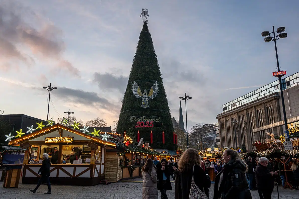 In der Abenddämmerung herrscht auf einem festlichen Weihnachtsmarkt unter freiem Himmel reges Treiben. In der Mitte steht ein riesiger, mit Lichtern, einem Engel und dem Schild "Dortmund 2025" geschmückter Weihnachtsbaum, umgeben von mit Lichterketten beleuchteten Essens- und Geschenkeständen.