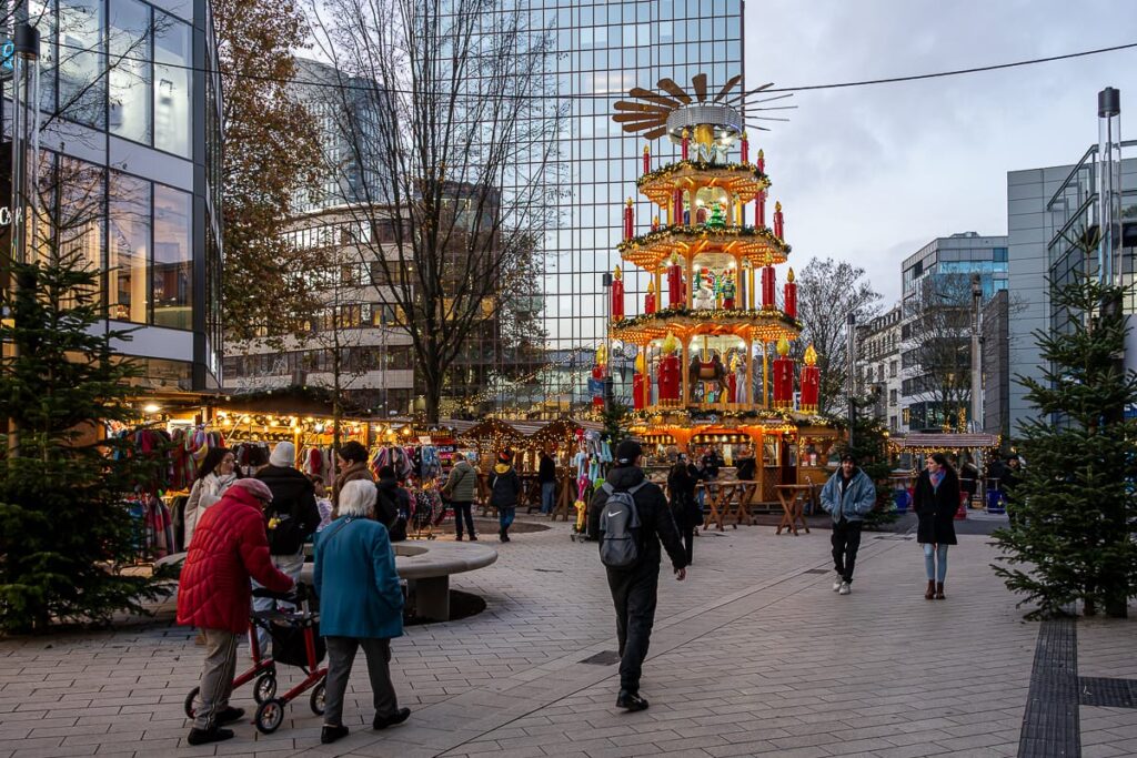 Eine festliche Weihnachtsmarktszene auf einem Stadtplatz zeigt ein hohes, beleuchtetes Holzkarussell mit Kerzen, Figuren und einem leuchtenden Weihnachtsbaum. Menschen gehen spazieren und kaufen ein; moderne Glastürme umgeben den lebhaften, fröhlichen Markt.