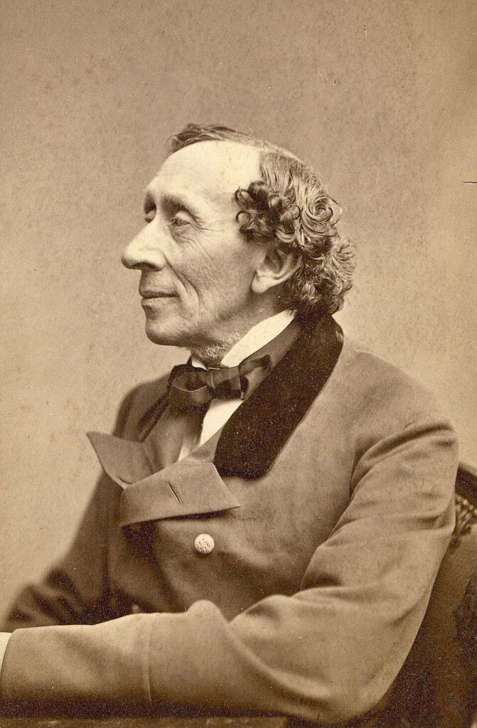 Sepia getöntes Porträt von Hans Christian Andersen im Profil, mit gewelltem Haar, markanter Nase und leichtem Lächeln. Er trägt einen formellen Mantel mit Samtkragen, Fliege und Zierknopf und sitzt mit verschränkten Armen auf einem gemusterten Stuhl.