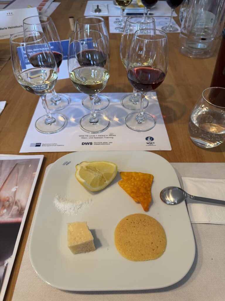 Ein Verkostungsset der WSET-Stufe 1 mit sechs Weingläsern, Wasser und einem Teller mit Zitrone, Salz, Hartkäse, einem gelben Cracker und einem orangefarbenen Tortilla-Chip. Ein Löffel und eine Serviette liegen neben dem Teller, und unter den Gläsern liegen Papiere.