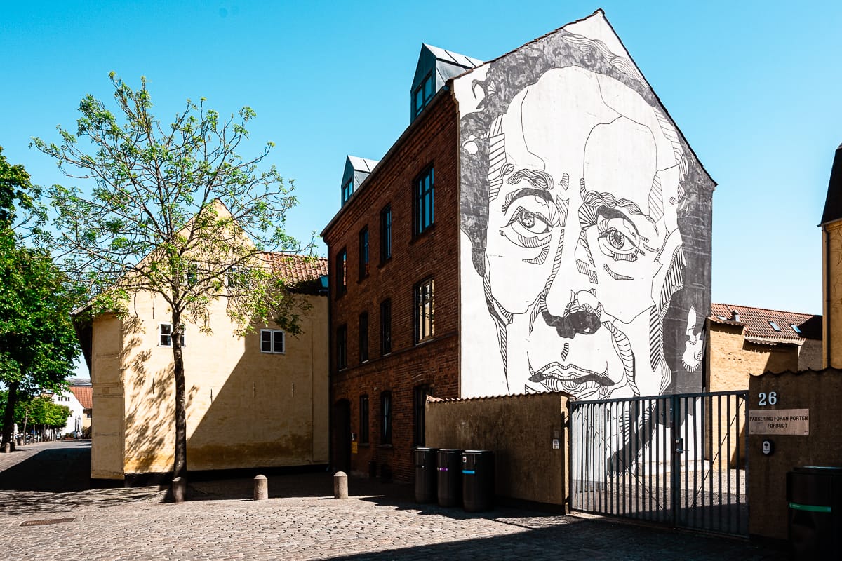 Ein großes, detailliertes Wandgemälde mit dem Gesicht von Hans Christian Andersen ist auf die Seite eines Backsteingebäudes gemalt. Das Gebäude steht auf einer Kopfsteinpflasterstraße mit einem Baum in der Nähe unter einem klaren blauen Himmel, gelben Gebäuden und einem Metalltor im Blick.