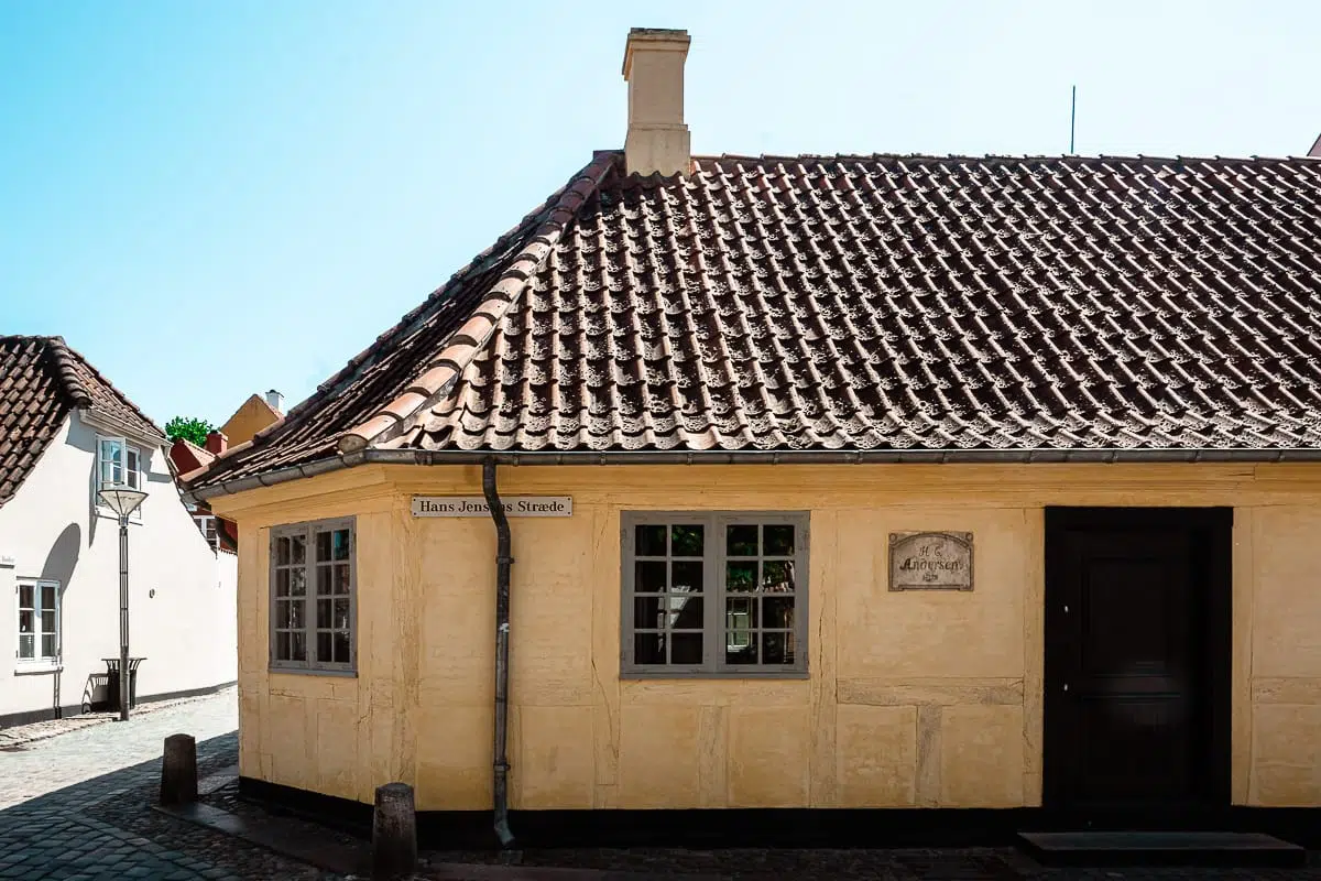 Ein kleines, gelbes, historisches Haus mit rotem Ziegeldach steht an einer kopfsteingepflasterten Straßenecke namens "Hans Jensen Stræde". In der Nähe befinden sich verschlossene Fenster und eine hölzerne Eingangstür. Auf einem Schild steht H.C. Andersens Hus.