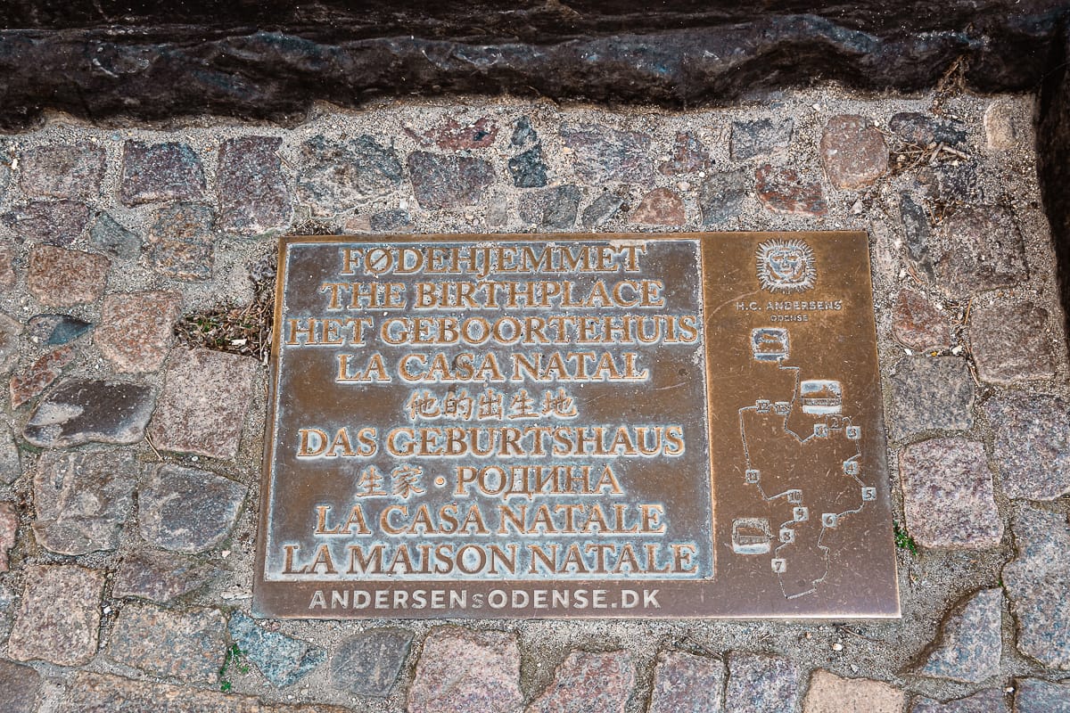 Eine in Kopfsteinpflaster eingelassene Bronzetafel markiert das Geburtshaus von Hans Christian Andersen. "Das Geburtshaus" ist in neun Sprachen beschriftet, mit dekorativen Motiven, der Website andersensodense.dk und Anzeichen von Patina durch Alter und Witterung.