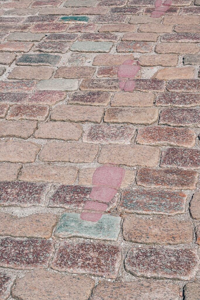 Ein Kopfsteinpflasterweg mit rosafarbenen Fußabdrücken, von Hans Christian Andersen, führt diagonal von der unteren Mitte nach oben links. Einige Steine sind hellgrau, die meisten rötlich-braun, und die Sonne beleuchtet die Szene im Freien hell.