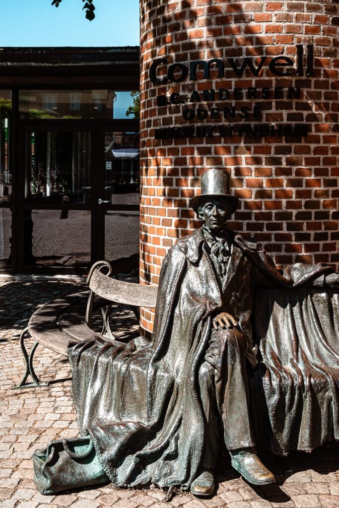 Eine Bronzestatue von Hans Christian Andersen mit Zylinder und Mantel sitzt auf einer Bank neben einer runden Backsteinmauer mit der Aufschrift "Comwell Andersen Odense". Das Sonnenlicht wirft Schatten auf den kopfsteingepflasterten Boden und die spiegelnden Glasfenster dahinter.