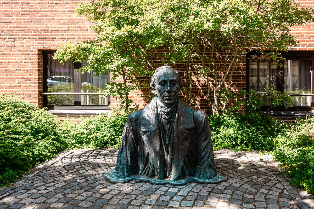 Eine Bronzebüste zeigt den oberen Teil von  Hans Christian Andersen in einem Anzug. Er scheint aus dem Boden heraus zu kommen. Statue steht im Freien auf Kopfsteinpflaster. Grüne Büsche und ein belaubter Baum umgeben die Statue. Ein Backsteingebäude und seine Fenster sind durch das Laub teilweise sichtbar.
