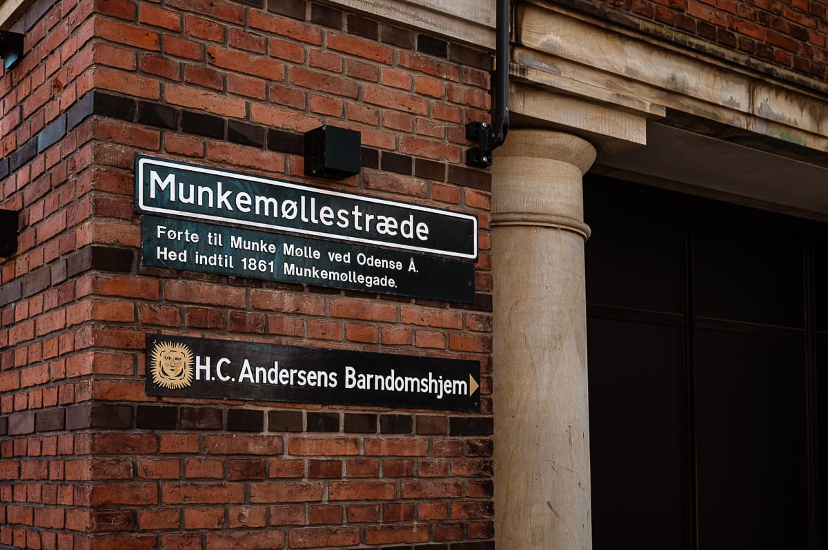 Eine Backsteinmauer mit drei Schildern: "Munkemøllestræde", eine dänische Beschreibung über Munke Mølle und Odense, und "H.C. Andersens Barndomshjem" mit einem goldenen Emblem, das das Elternhaus von Hans Christian Andersen kennzeichnet.