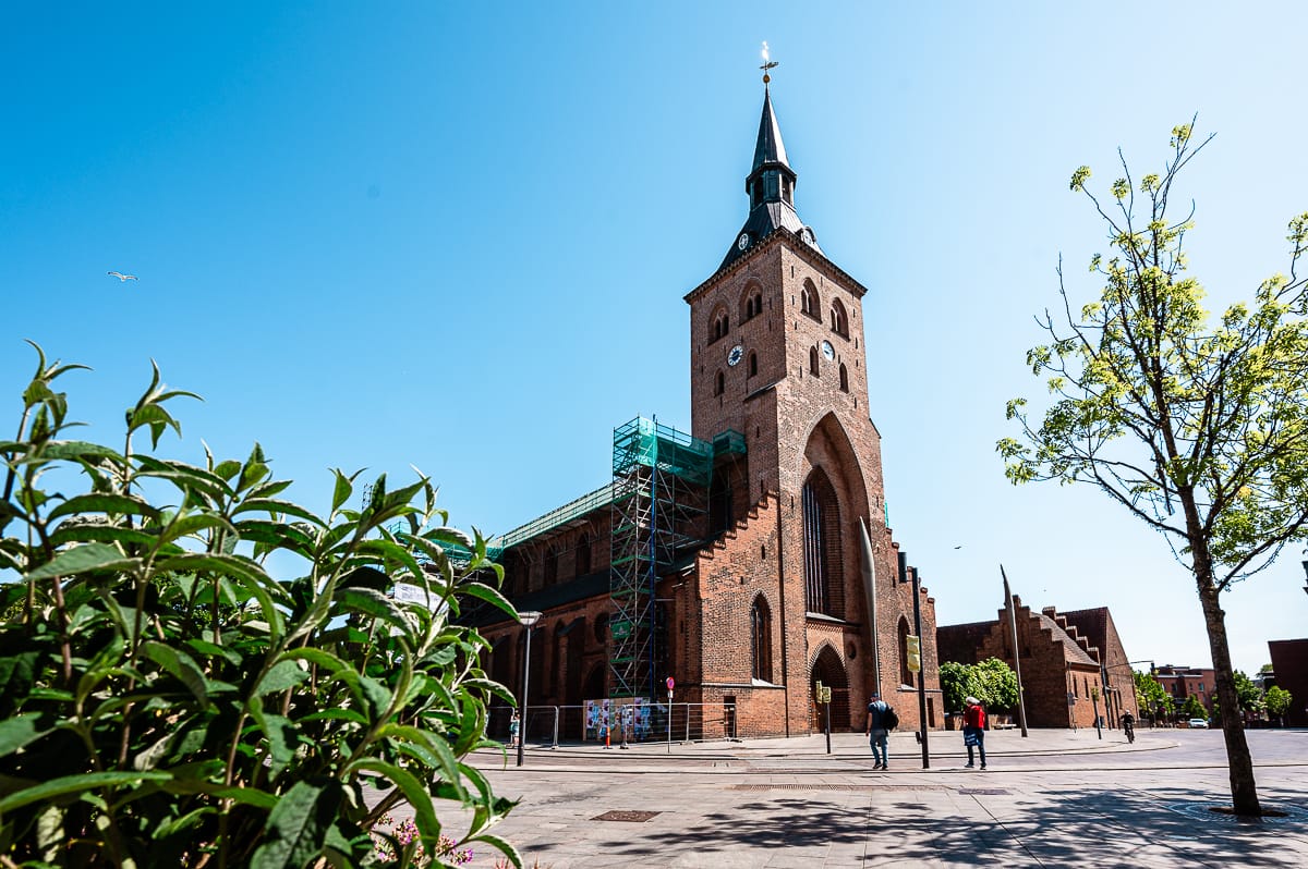 Eine große Backsteinkirche mit einem hohen Glockenturm und einer Turmspitze steht unter einem klaren blauen Himmel. Ein Gerüst bedeckt einen Teil des Gebäudes. In der Nähe umrahmen grüne Pflanzen und ein Baum den sonnigen, offenen Platz.