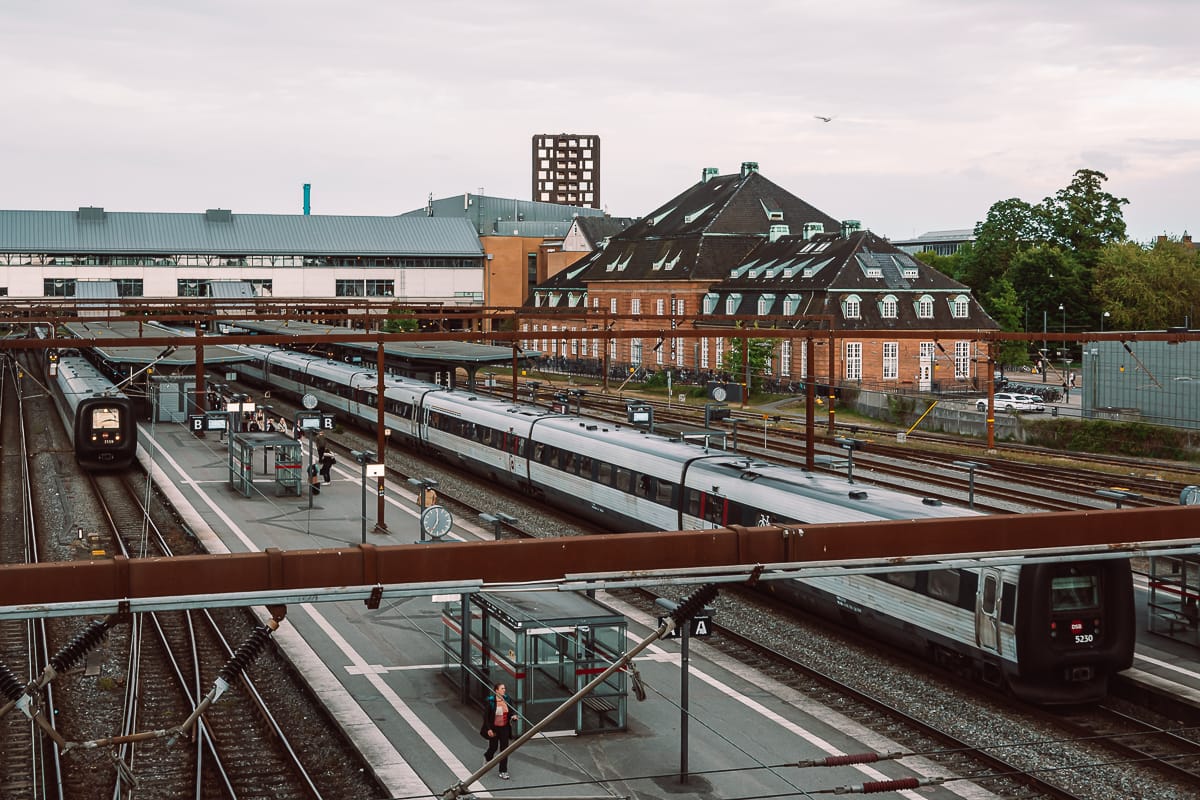 Ein Bahnhof mit mehreren Gleisen und Bahnsteigen, mehreren modernen Zügen, die geparkt sind, und einigen wartenden Fahrgästen. Darüber kreuzen sich Oberleitungen. Im Hintergrund das alte Bahnhof-Backsteingebäude.