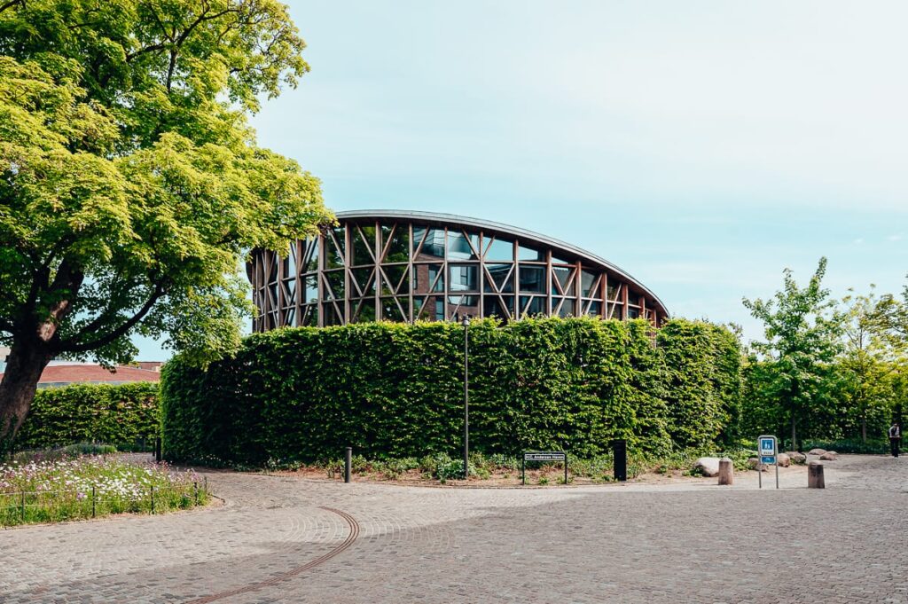 Ein modernes rundes Gebäude mit einer hölzernen Gitterfassade, die teilweise von grünem Efeu bedeckt ist, ist von Kopfsteinpflaster, Bäumen und einer Bank umgeben. 