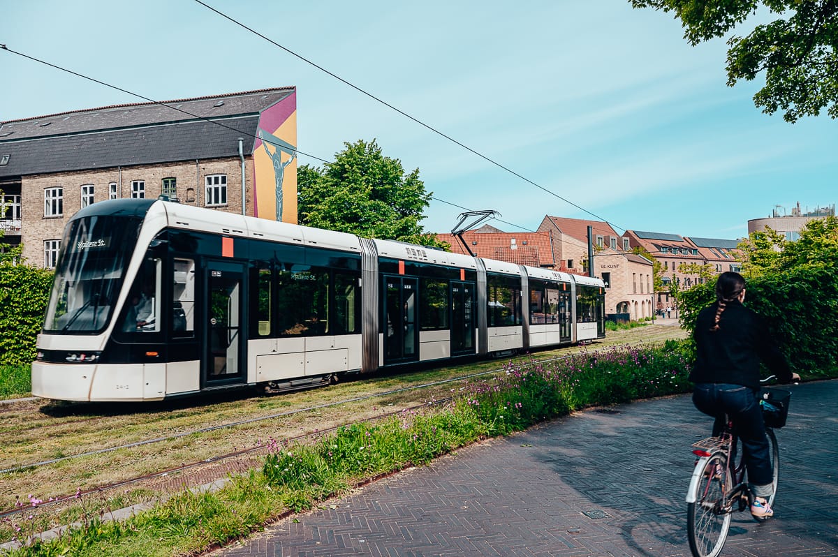 Eine moderne weiße Straßenbahn fährt auf grasbewachsenen Gleisen neben einem Backsteingebäude mit einer bunten Wandmalerei. Im Vordergrund tritt eine Person mit Pferdeschwanz in die Pedale unter dem klaren blauen Himmel.