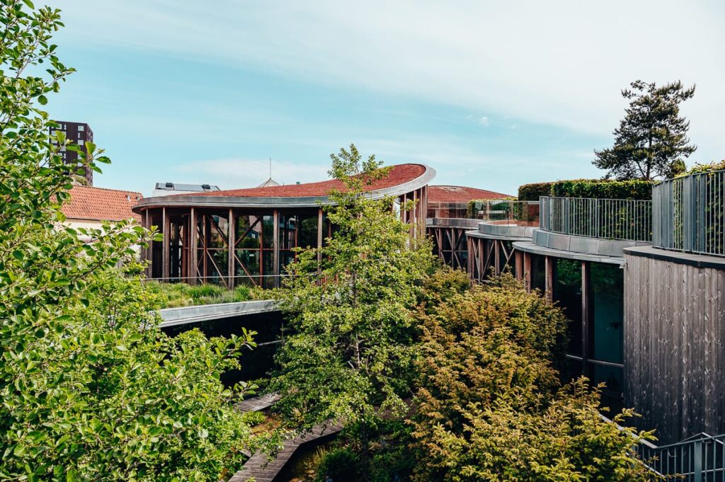 Ein modernes Gebäude mit großen Glasfenstern und einem roten geschwungenen Dach ist von üppig grünen Bäumen umgeben und zeigt einen Teil des unterirdischen Hans Christian Andersen Museum. Ein erhöhter Gehweg schlängelt sich durch die Landschaft und lässt Architektur und Natur unter blauem Himmel miteinander verschmelzen.