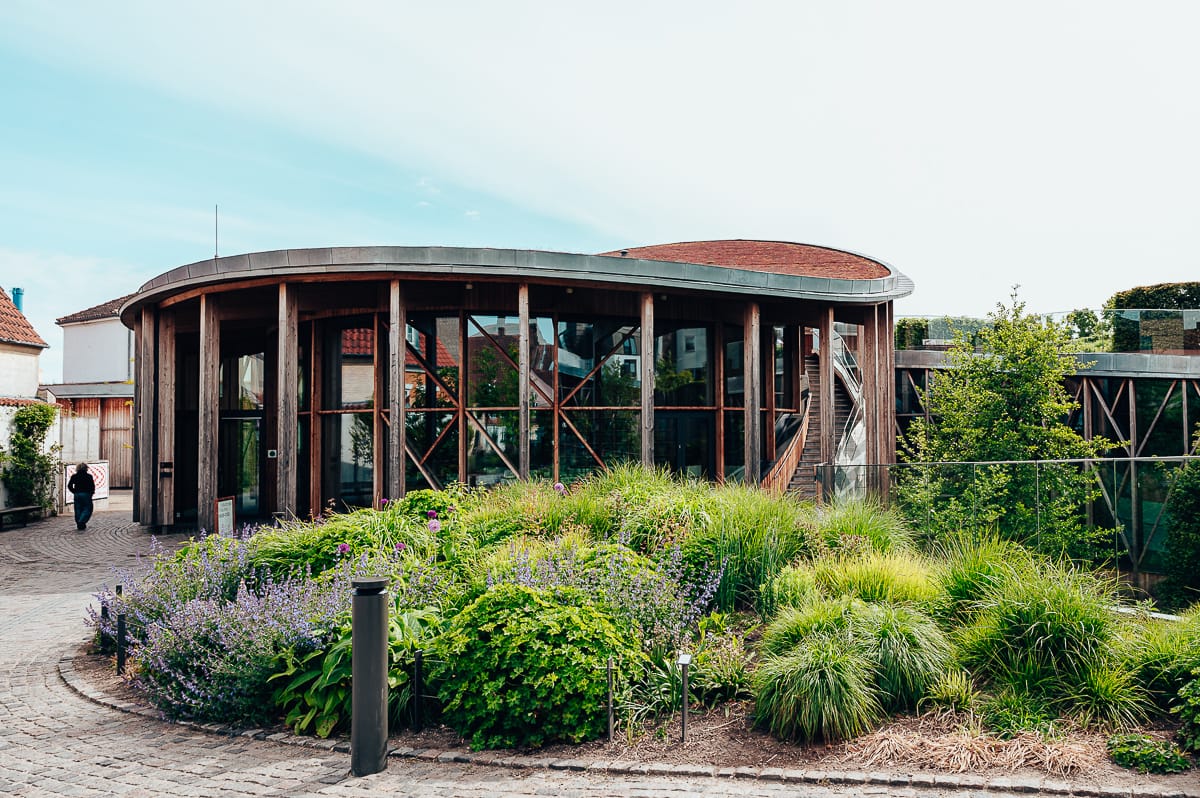 Hinter einem üppigen Garten mit grünen Sträuchern und violetten Blumen steht ein modernes Gebäude mit großen Glasfenstern und geschwungenen Holzbalken, der Eingang des Hans Christian Andersen Museums. Ein kopfsteingepflasterter Weg führt unter einem hellen, teilweise bewölkten Himmel zum Eingang.
