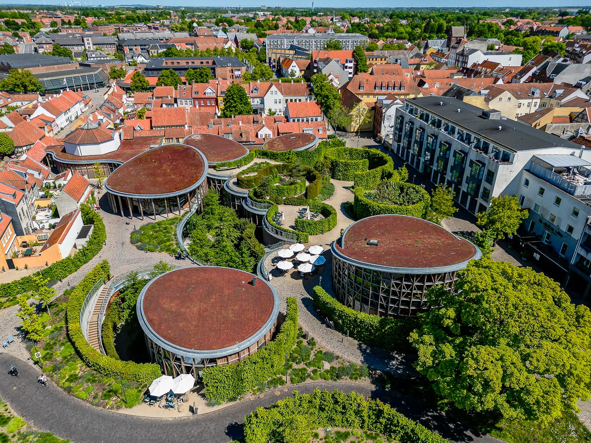 Luftaufnahme einer Stadtlandschaft mit einzigartigen, runden Gebäuden mit roten Dächern, die von Grünflächen, Höfen und Wegen umgeben sind. Traditionelle Häuser mit orangefarbenen Dächern stehen neben modernen Gebäuden unter einem klaren Himmel.