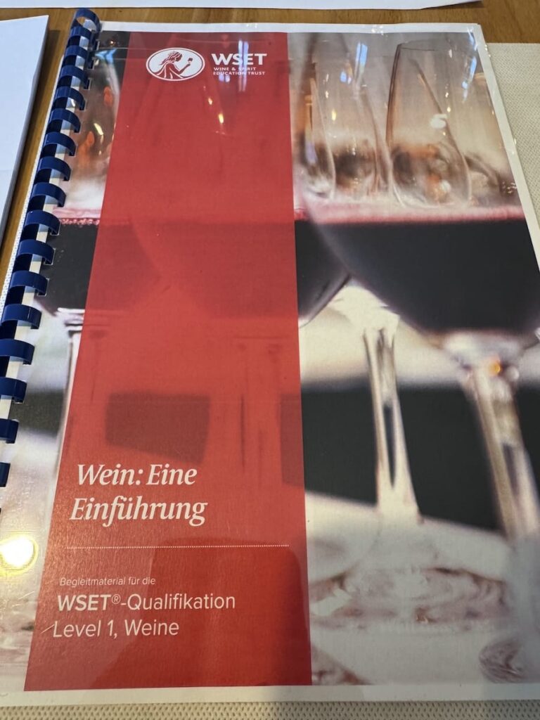 Ein spiralgebundenes Büchlein mit dem Titel "Wein: Eine Einführung" liegt auf einem Tisch. Auf dem rot-weißen Einband sind das WSET-Logo und Weingläser mit Rotwein abgebildet, was es als offizielles Studienbuch für die WSET Level 1 Wine Qualification in deutscher Sprache ausweist.
