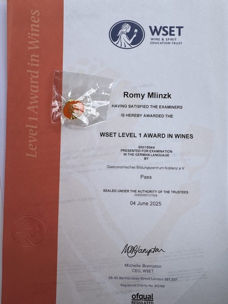 Ein WSET Level 1 Award in Wines-Zertifikat für Romy Mlinzk liegt auf einer flachen Oberfläche. Das WSET Level 1-Zertifikat ist mit einem roten Band, einem WSET-Siegel/Prägung, offiziellen Unterschriften und eine ovale Anstecknadel mit dem Traubenlogo des WSET liegt auf dem Papier.