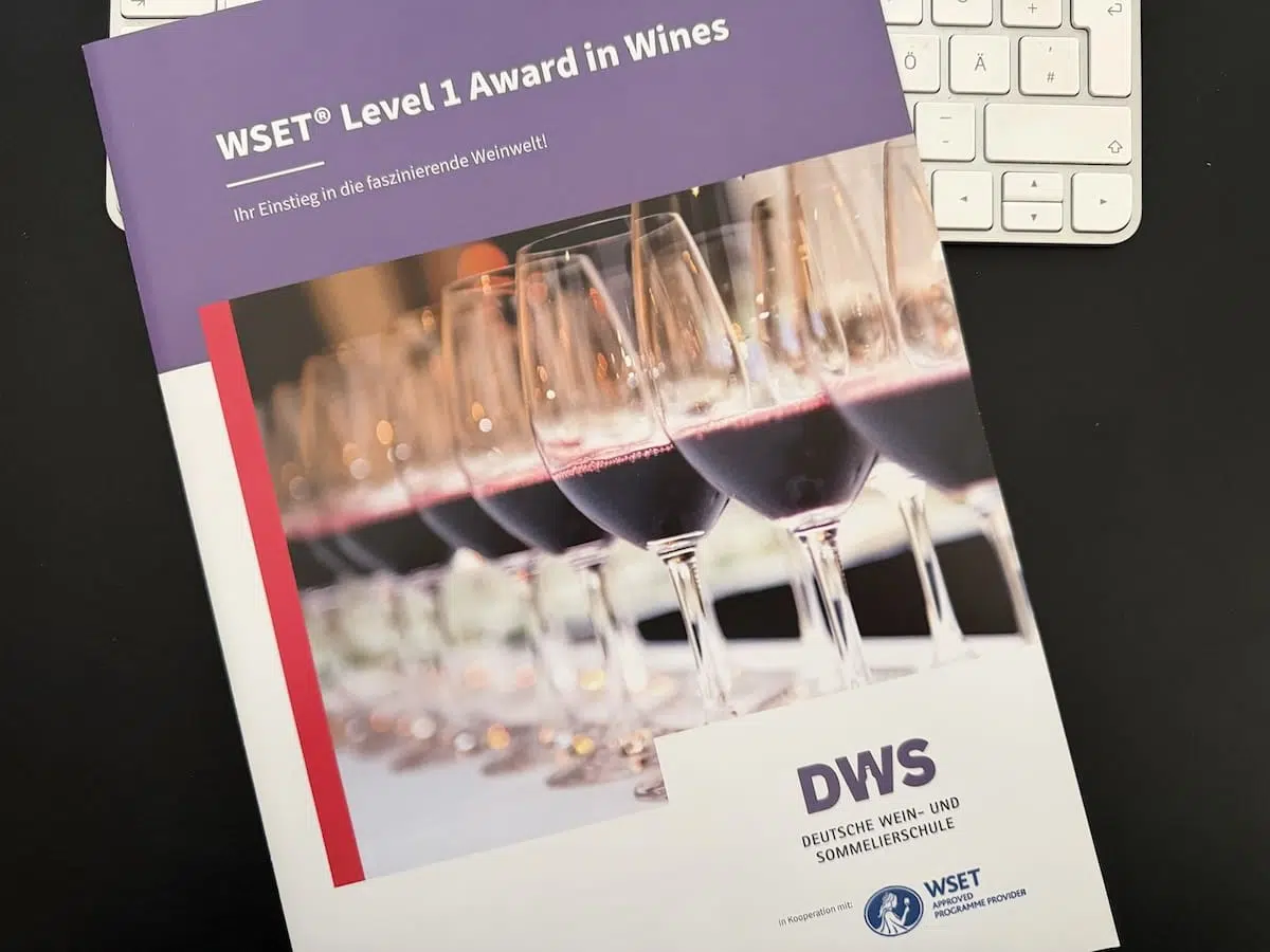 Eine Broschüre mit dem Titel "WSET® Level 1 Award in Wines" liegt auf einer schwarzen Fläche und verdeckt teilweise eine weiße Tastatur. Auf dem Umschlag sind mit Rotwein gefüllte Weingläser zu sehen, darunter das Logo der DWS (Deutsche Wein- und Sommelierschule) und das WSET®-Logo.