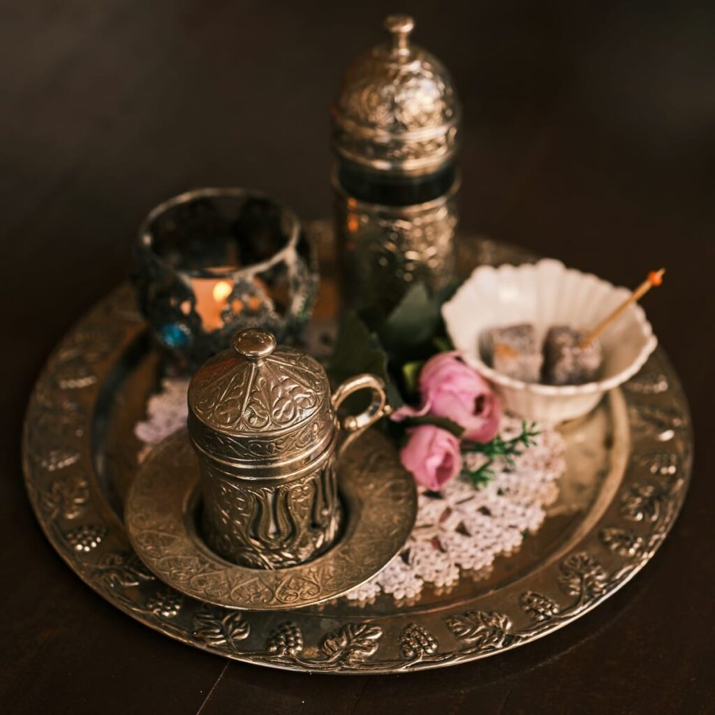 Ein dekoratives Metalltablett enthält eine verzierte Kaffeetasse mit Deckel, ein passendes hohes Gefäß, einen Kerzenhalter aus Glas, eine Schale mit türkischen Köstlichkeiten für den Ramadan und rosa Rosen auf einem gehäkelten Deckchen, alles auf einer dunklen Holzfläche.