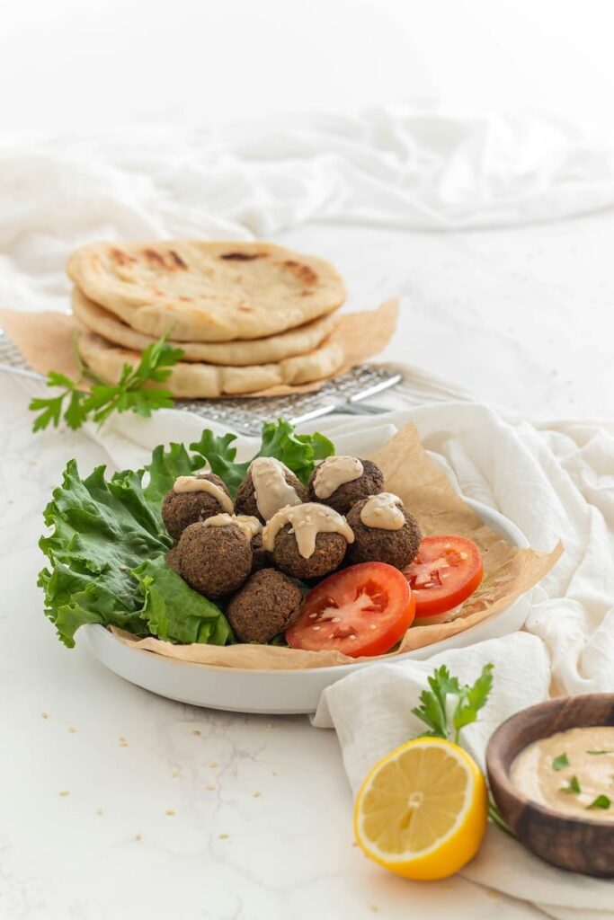 Ein Teller mit Falafelbällchen, die mit Tahinisauce beträufelt sind, liegt auf einem Salat mit Tomatenscheiben - perfekt für ein Ramadan-Essen. Daneben liegen Fladenbrote, eine Schale mit Soße, eine Zitronenhälfte und Petersilie, alles auf einer weißen, marmorartigen Fläche.