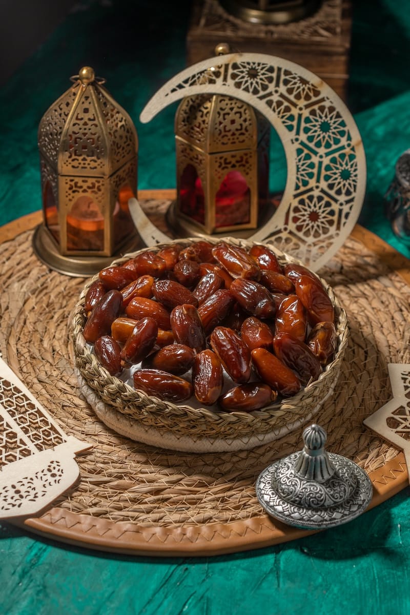 Ein geflochtener Korb, gefüllt mit Datteln, steht auf einer runden Strohmatte. Zwei verschnörkelte Laternen, eine dekorative Mondsichel und kunstvolle Scherenschnitte vermitteln eine festliche Ramadan-Atmosphäre, und ein silberner Behälter vervollständigt die Szene auf einer grünen Fläche.