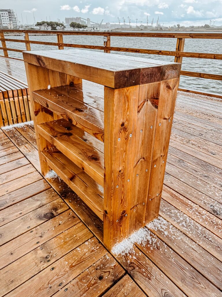 Ein Holzregal steht auf einem nassen Steg in der Nähe des Wassers in Landskrona, auf dem Hagelkörner verstreut sind. Das Regal hat drei offene Fächer, die aus dicken, polierten Holzbrettern bestehen. Auf der anderen Seite des Wassers sind Gebäude und Kräne zu sehen.
