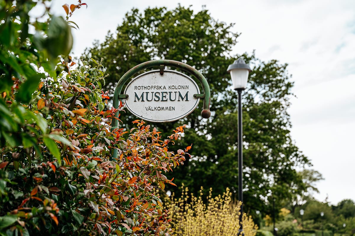 Ein altes Metallschild mit der Aufschrift "Rothofska Kolonin Museum Välkommen" hängt inmitten von grünem und rotem Laub in Landskrona. Laubbäume und eine alte Straßenlaterne stehen unter einem hellen, teilweise bewölkten Himmel und schaffen eine einladende und üppige Szene.