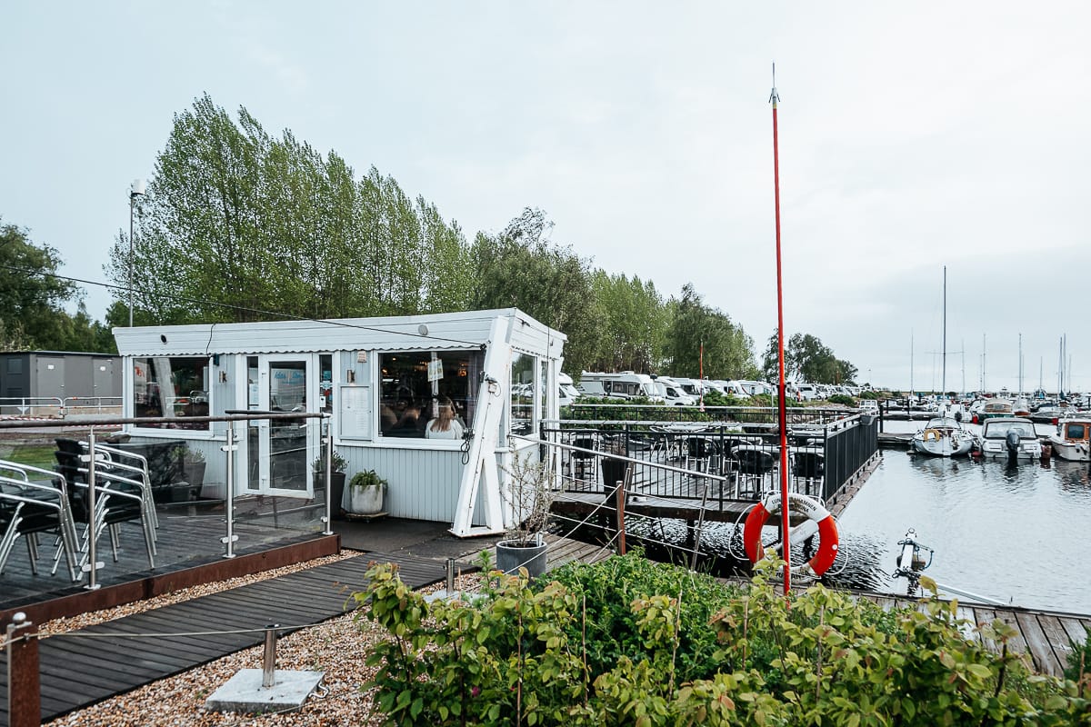 Ein kleines weißes Café mit großen Fenstern liegt auf einem Steg am Yachthafen von Landskrona. Die Sitzgelegenheiten im Freien mit Metallstühlen und -tischen stehen auf einem Holzdeck mit Blick auf die im Wasser vertäuten Boote. Im Hintergrund stehen Bäume und geparkte Fahrzeuge unter einem wolkenverhangenen Himmel.