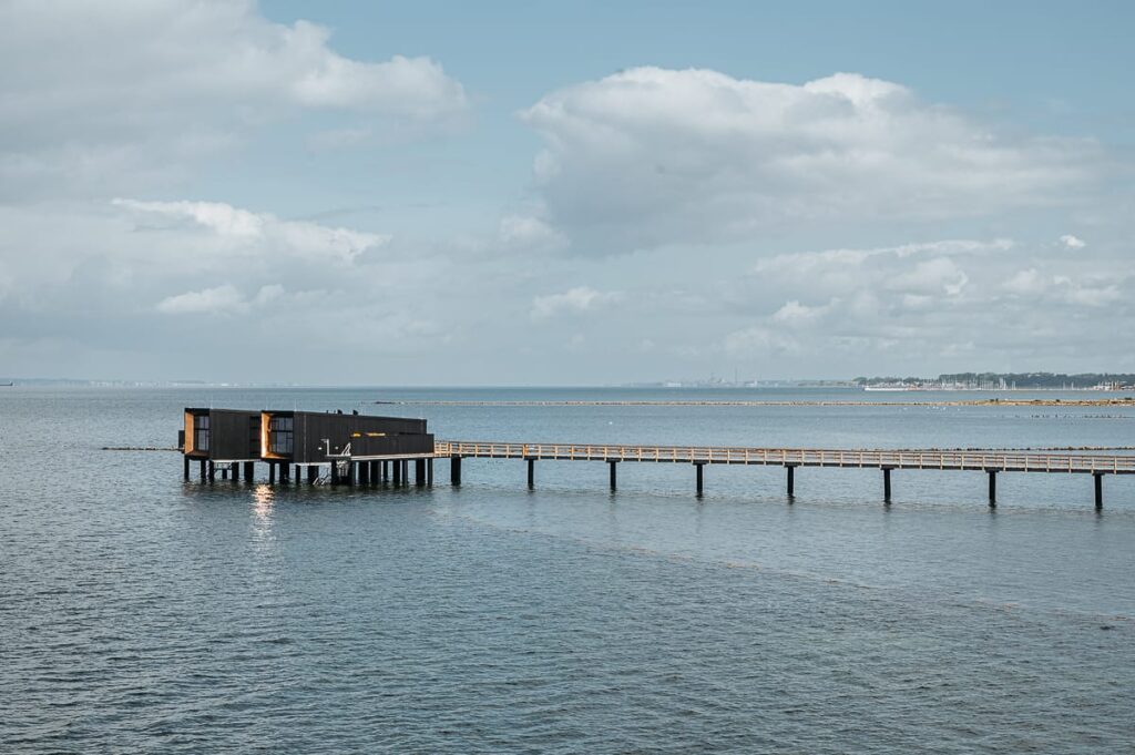 Ein modernes schwarzes Gebäude auf Stelzen ragt über das ruhige blaue Wasser in Landskrona, das durch eine lange Holzpromenade mit dem Ufer verbunden ist. Der Himmel ist teilweise bewölkt, und das ferne Land erscheint am Horizont in einem weichen Licht.