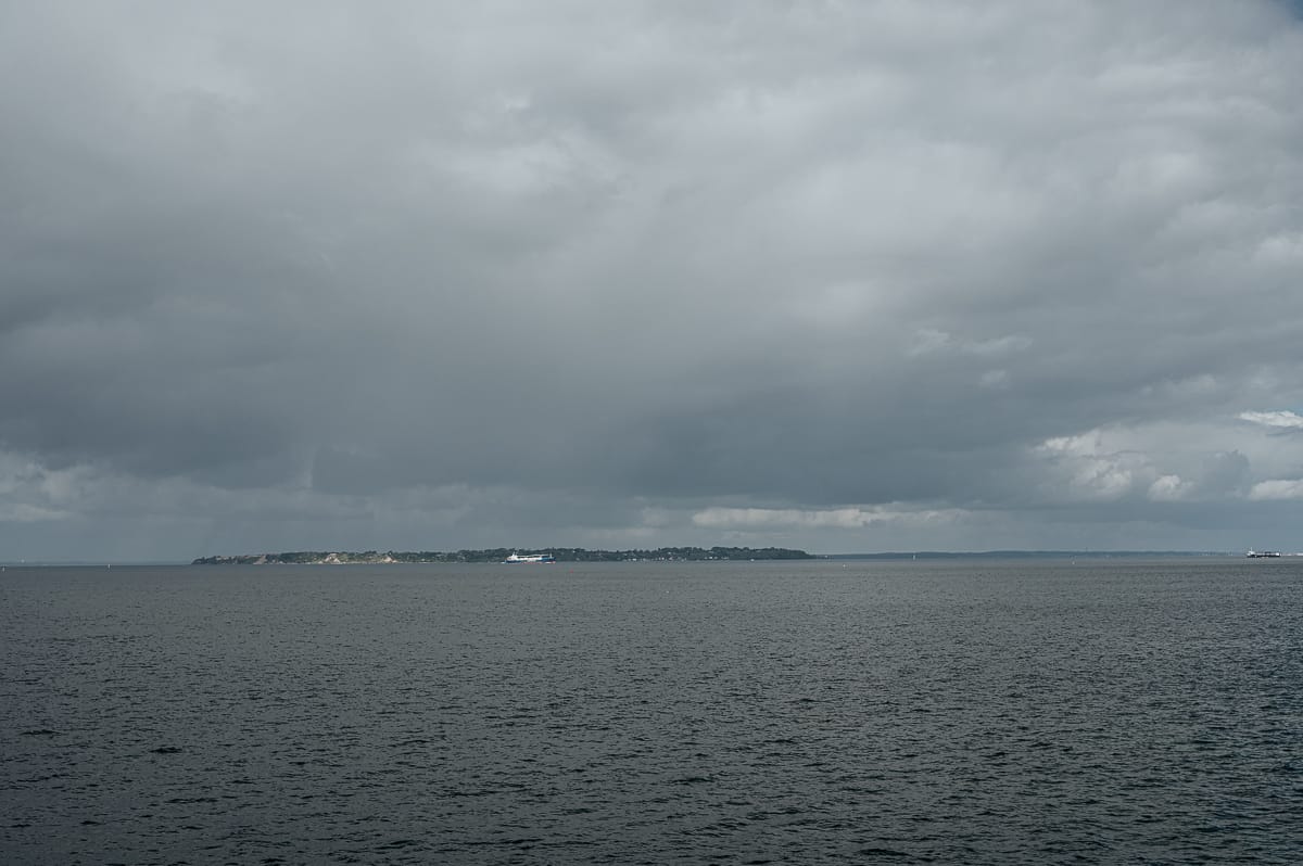 Eine entfernte, niedrig gelegene Insel in der Nähe von Landskrona erstreckt sich am Horizont unter einem bewölkten, bedeckten Himmel. Das ruhige Meer im Vordergrund erscheint dunkelgrau, was das gedämpfte Licht widerspiegelt und mit gedämpften Farben ein ruhiges Gefühl der Isolation hervorruft.