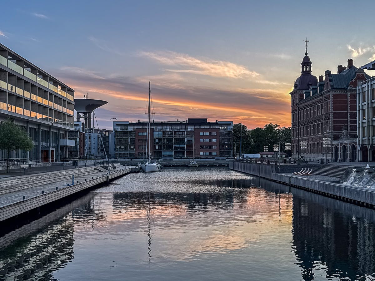 In einem ruhigen Kanal in Landskrona spiegeln sich moderne Wohnungen und ein historisches Gebäude aus rotem Backstein mit einer Kuppel und einem Turm. Am Ende des Kanals liegen zwei Segelboote vor Anker, und der pastellfarbene Abendhimmel spiegelt sich deutlich im ruhigen Wasser wider.