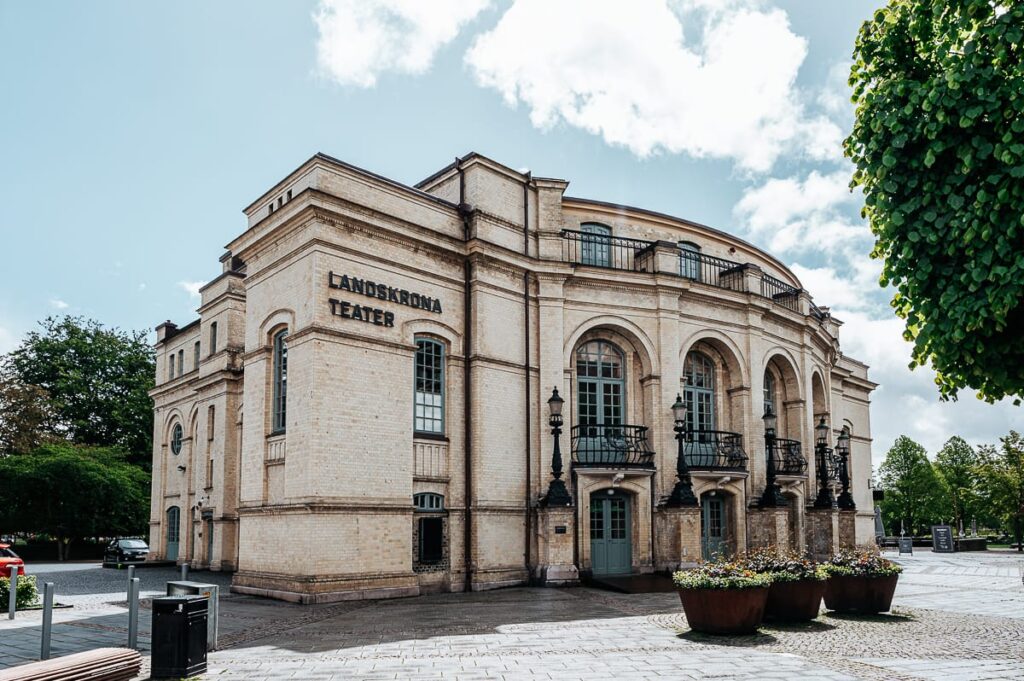 Das historische Landskrona Teater ist ein prächtiges, helles Backsteingebäude mit Bogenfenstern, Säulen und schwarzen Geländern. Umgeben von Bäumen, Bänken und einer Straße mit Kopfsteinpflaster ziert es das Herz von Landskrona unter einem teilweise bewölkten Himmel.