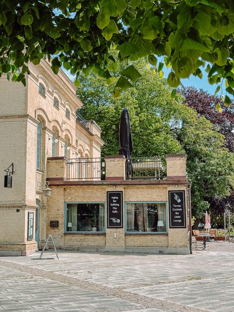 Ein helles Backsteingebäude mit gewölbten Fenstern und einer Terrasse liegt an einem ruhigen Platz. Schilder werben für ein Café und eine Lounge. Grüne Bäume rahmen das Bild ein, und im Hintergrund sind Sitzplätze im Freien unter einem klaren, hellen Himmel zu sehen.