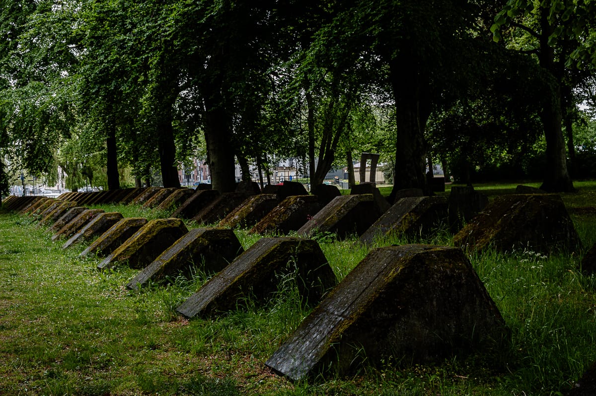 Eine Reihe großer, moosbewachsener, pyramidenförmiger Betonblöcke steht auf einer Wiese unter hohen, belaubten Bäumen in Landskrona. Die Szenerie wirkt schattig und ruhig, da das Sonnenlicht durch das dichte Blattwerk fällt.