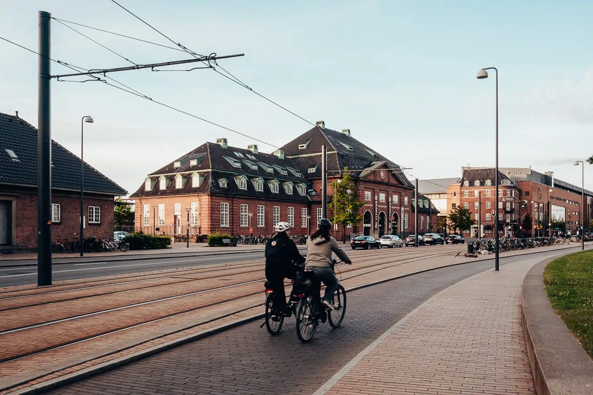 Zwei Personen fahren mit dem Fahrrad auf einem gepflasterten Weg neben den Straßenbahnschienen in Odense, einer Stadt mit klassischen Backsteinbauten. Der Himmel ist meist klar, und die Straßenlaternen säumen die Straße, was der ruhigen städtischen Szene ein frühabendliches oder spätnachmittägliches Licht verleiht.