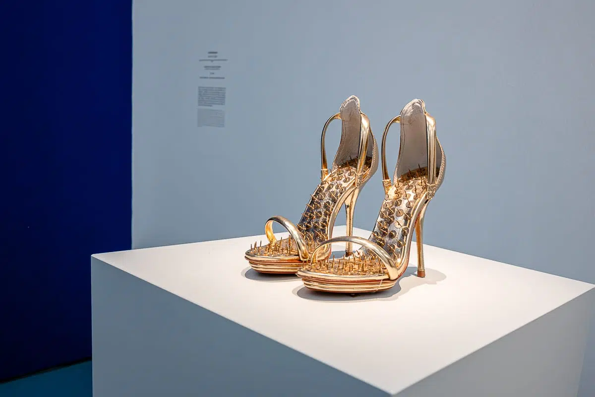 Ein Paar hochhackige goldene Sandalen mit Riemen sind auf einem weißen Sockel in einer Galerie ausgestellt, vor einer weiß-blauen Wand mit einer verschwommenen Texttafel im Hintergrund. Auf dem Fußbett sind lauter goldene Reißzwecken mit der Nadel nach oben aufgebracht.