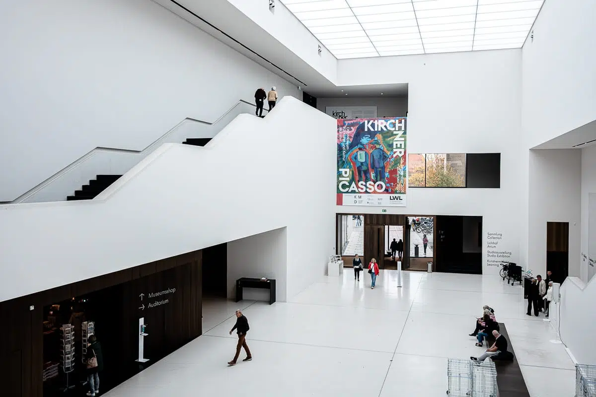 Eine geräumige, moderne Museumslobby mit weißen Wänden, hohen Decken und einem großen Oberlicht. Die Besucher gehen und sitzen auf Bänken. In der Nähe des Eingangs ist ein buntes Kunstplakat zur Ausstellung Kirchner.Picasso aufgehangen, und eine Treppe führt zu einer oberen Ebene, auf der zwei Personen stehen.