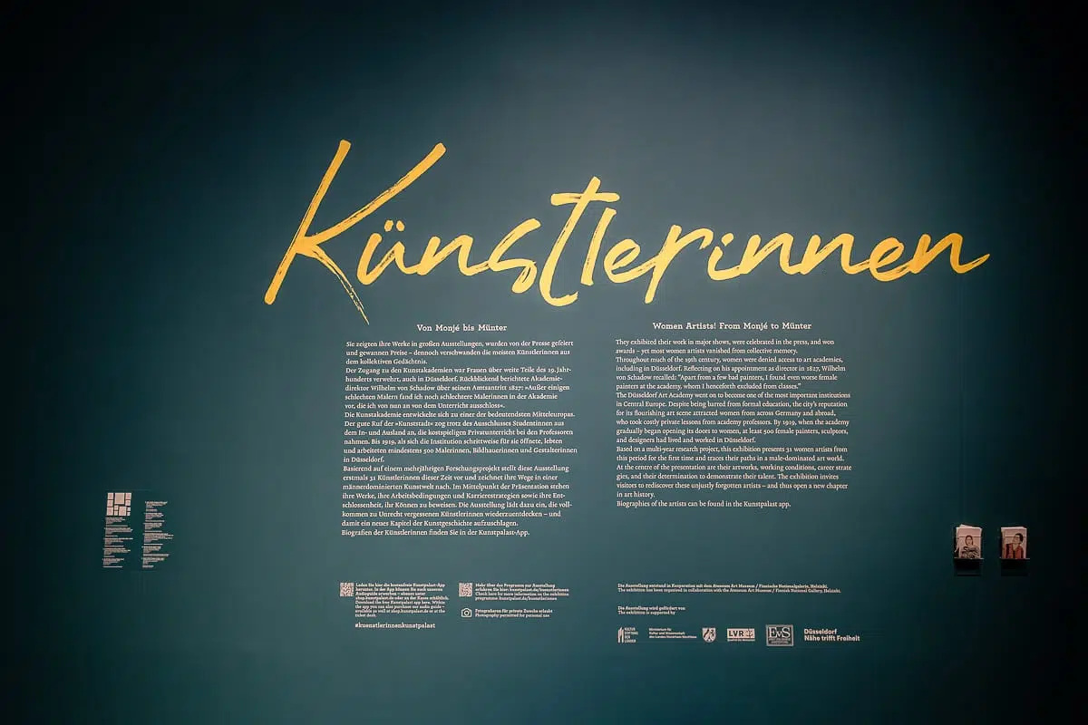 Auf einer dunkelgrünen Wand steht in großen gelben Lettern "Künstlerinnen" in Schreibschrift. Darunter befinden sich zwei Spalten mit weißem Text in deutscher Sprache, in denen die vorgestellten Künstlerinnen mit Logos und zwei Porträtfotos in einem Museum oder einer Galerie vorgestellt werden.