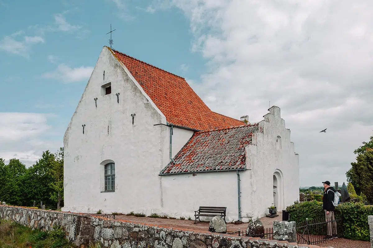 Eine kleine, weiß getünchte Steinkirche mit einem steilen roten Ziegeldach steht unter dem teilweise bewölkten Himmel von Ven. Eine Person mit einem Rucksack wartet am Eingang, in der Nähe eines Steinwegs, einer Bank und einer niedrigen Steinmauer, umgeben von üppigem Grün.