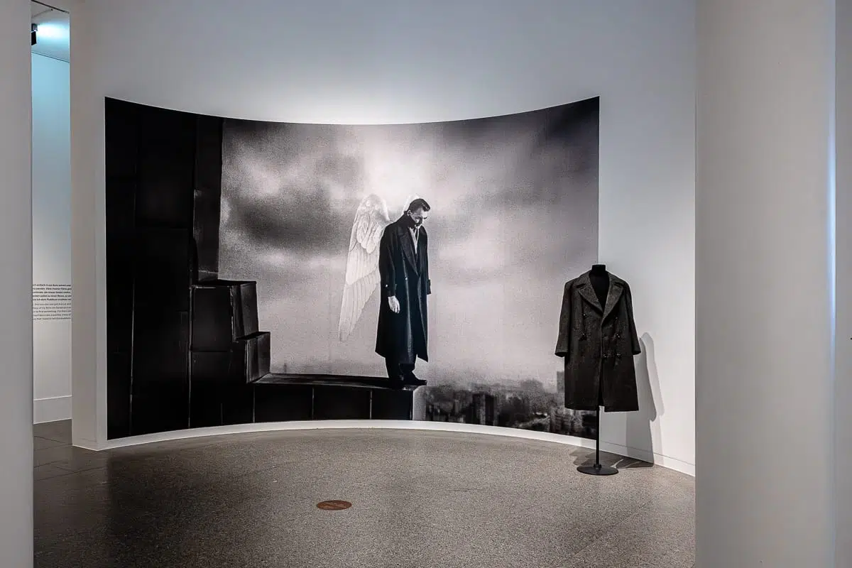 Ein Schwarz-Weiß-Foto aus dem Wim Wenders Film "Der Himmel über Berlin" zeigt einen Mann mit Engelsflügeln am Rande eines dunklen Gebäudes. Daneben ist ein langer schwarzer Mantel auf einer Schaufensterpuppe in einer sauberen, modernen Galerie mit weißen Wänden ausgestellt.