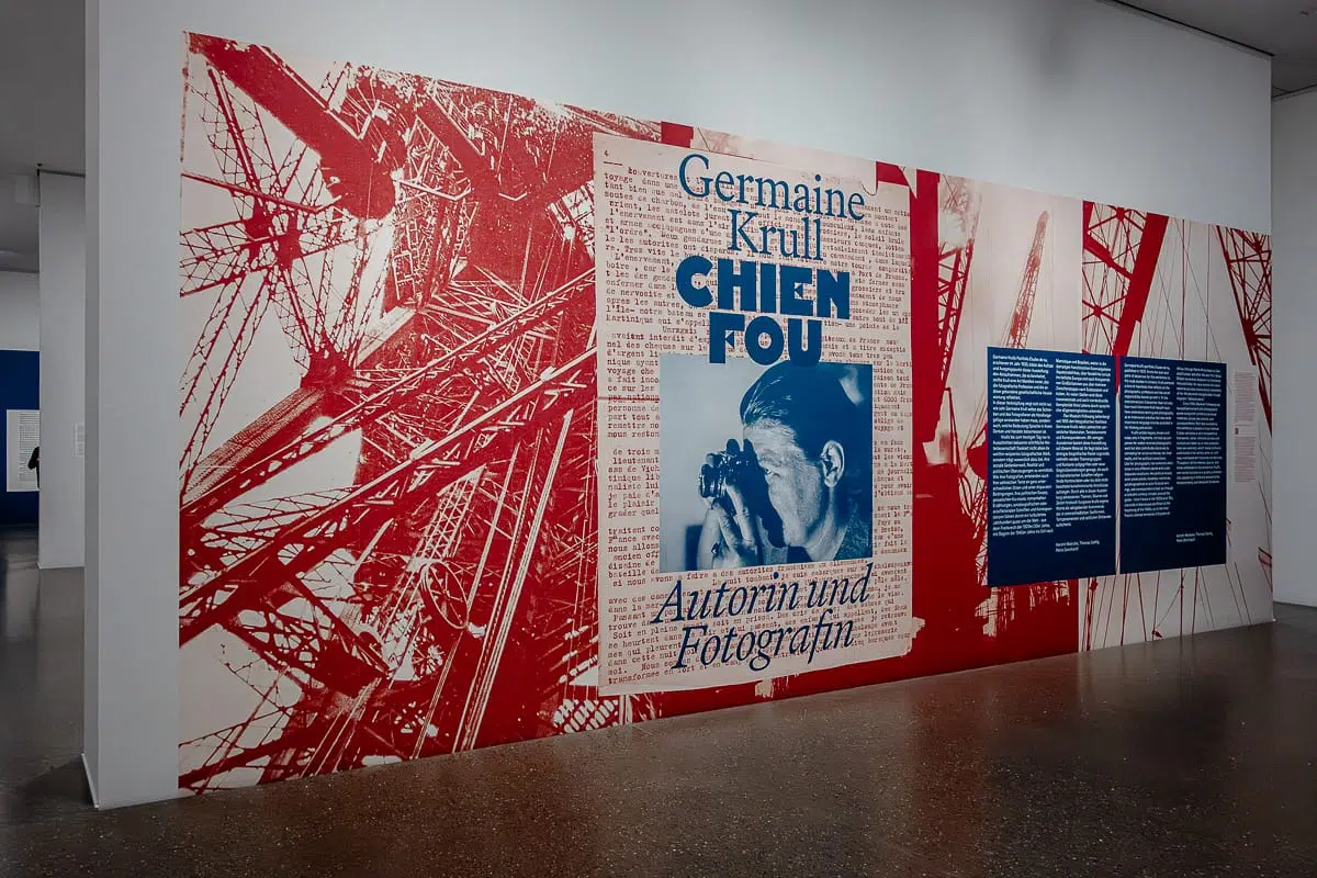 Eine Museumswand zeigt Germaine Krull mit einer auffälligen rot-weißen Industriegrafik, einem Porträt von ihr mit einer Kamera in der Hand und der fetten Aufschrift "Germaine Krull Chien Fou Autorin und Fotografin". Zwei blaue Tafeln mit weißem Text vervollständigen die Szene.