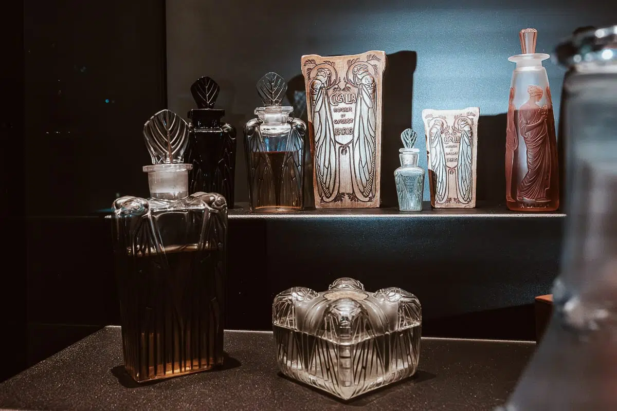 Eine Ausstellung von alten Glasparfümflaschen und verzierten Behältern, einige mit geätzten oder geformten Motiven im Musée Lalique. Dekorative Parfümverpackungen und ein hoher Milchglasflakon mit Stopfen leuchten in sanftem, konzentriertem Licht.