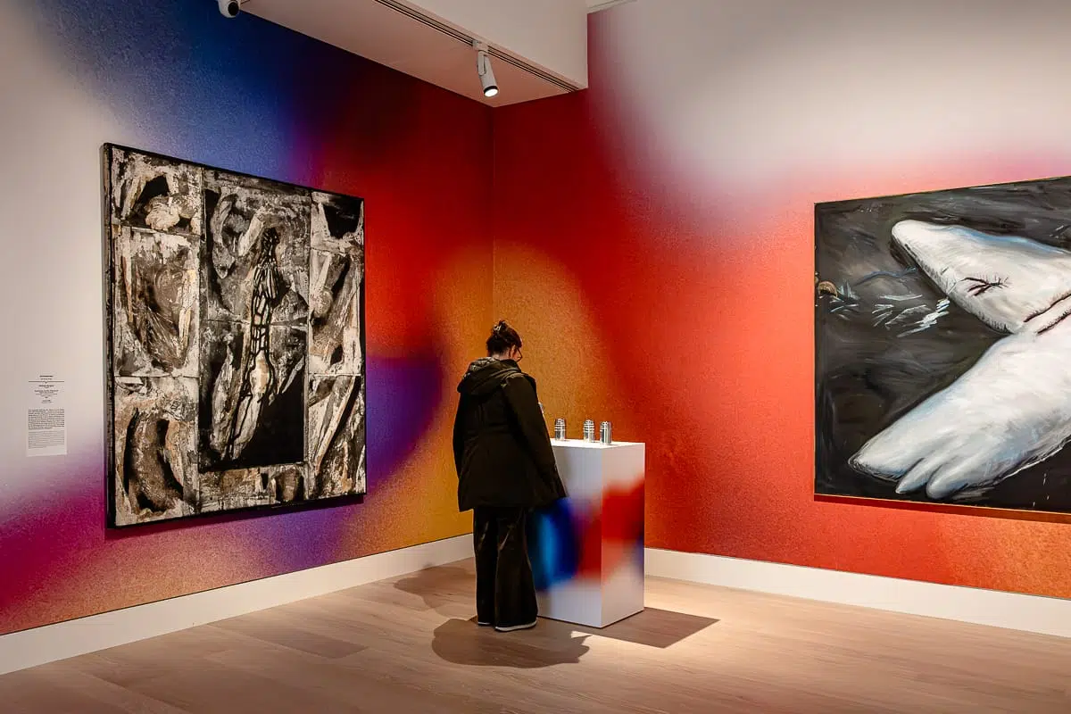 Eine Person steht in einer modernen Kunstgalerie und blickt auf ein kleines Ausstellungspodest. An den leuchtenden, bunten Wänden hängen abstrakte und surreale Gemälde - darunter ein Hai -, während subtile Düfte die Luft erfüllen. Die Galerie ist mit Holzböden und Scheinwerfern ausgestattet.