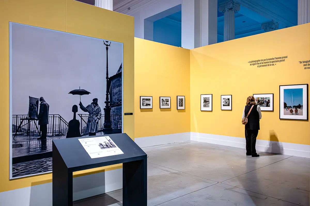 Eine Person betrachtet gerahmte Schwarzweiß- und Farbfotografien, darunter ein beeindruckendes Bild von Robert Doisneau, an gelben Galeriewänden. Ein großes Foto eines Künstlers, der im Freien im Regen malt, sticht zwischen hohen weißen Säulen und heller Museumsbeleuchtung hervor.
