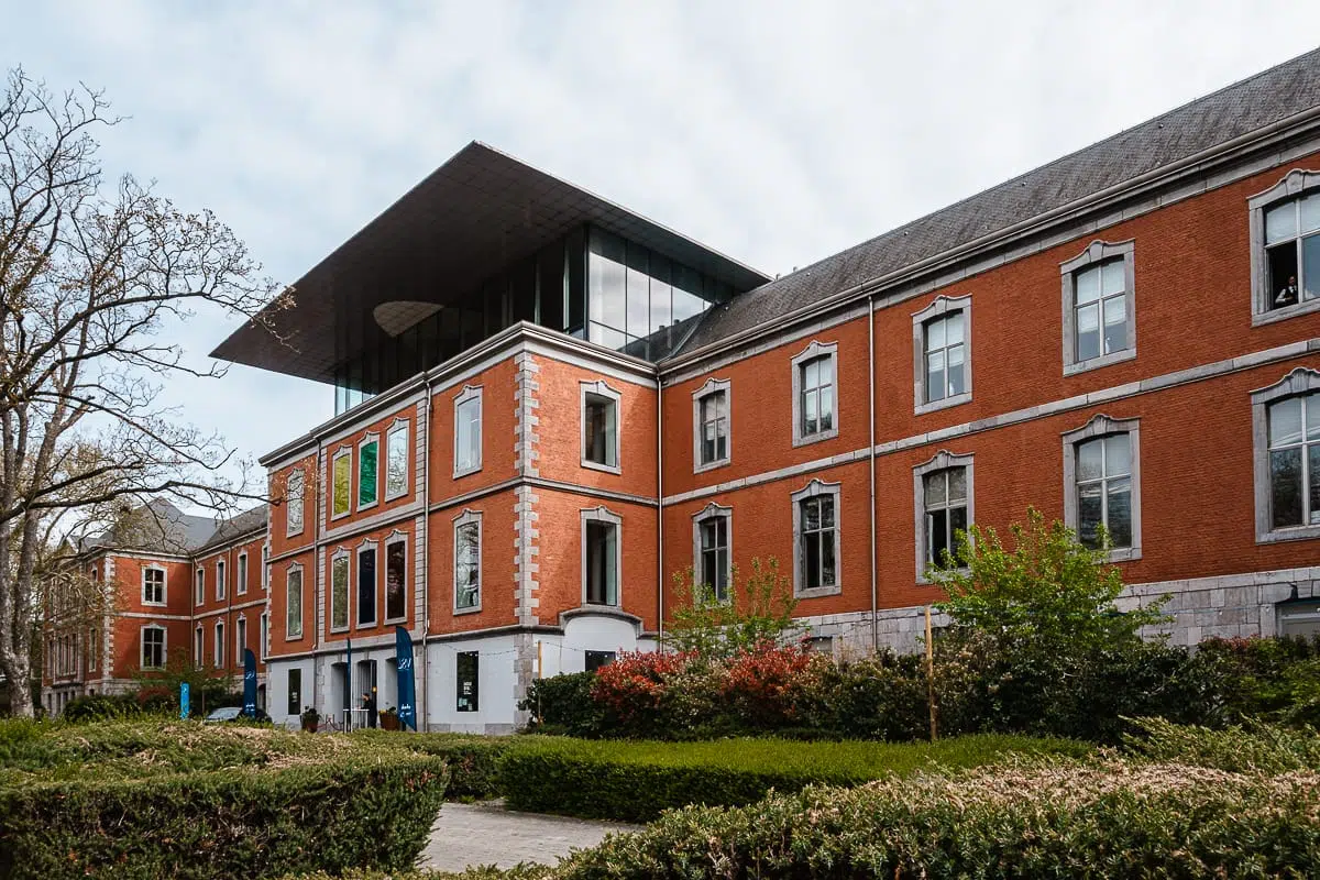 Das Gebäude aus rotem Backstein mit weißen Steinakzenten verfügt über einen modernen Glasanbau, der an die eleganten Entwürfe von Val Saint-Lambert erinnert. Das Gebäude ist von grünen Sträuchern und Bäumen umgeben, mit einem wolkenverhangenen Himmel darüber und mehreren Fenstern, die das Gebäude säumen.