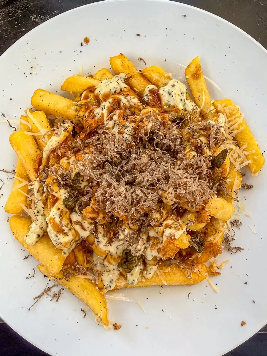Ein weißer Teller vom SchwarzGold, gefüllt mit dick geschnittenen Pommes frites, garniert mit cremiger Soße, geriebenem Käse, fein geriebener Trüffel und Kräutern. Die Pommes frites sind reichlich belegt, und die Garnierungen sind gleichmäßig auf dem Teller verteilt, was dem Gericht ein üppiges und abwechslungsreiches Aussehen verleiht.